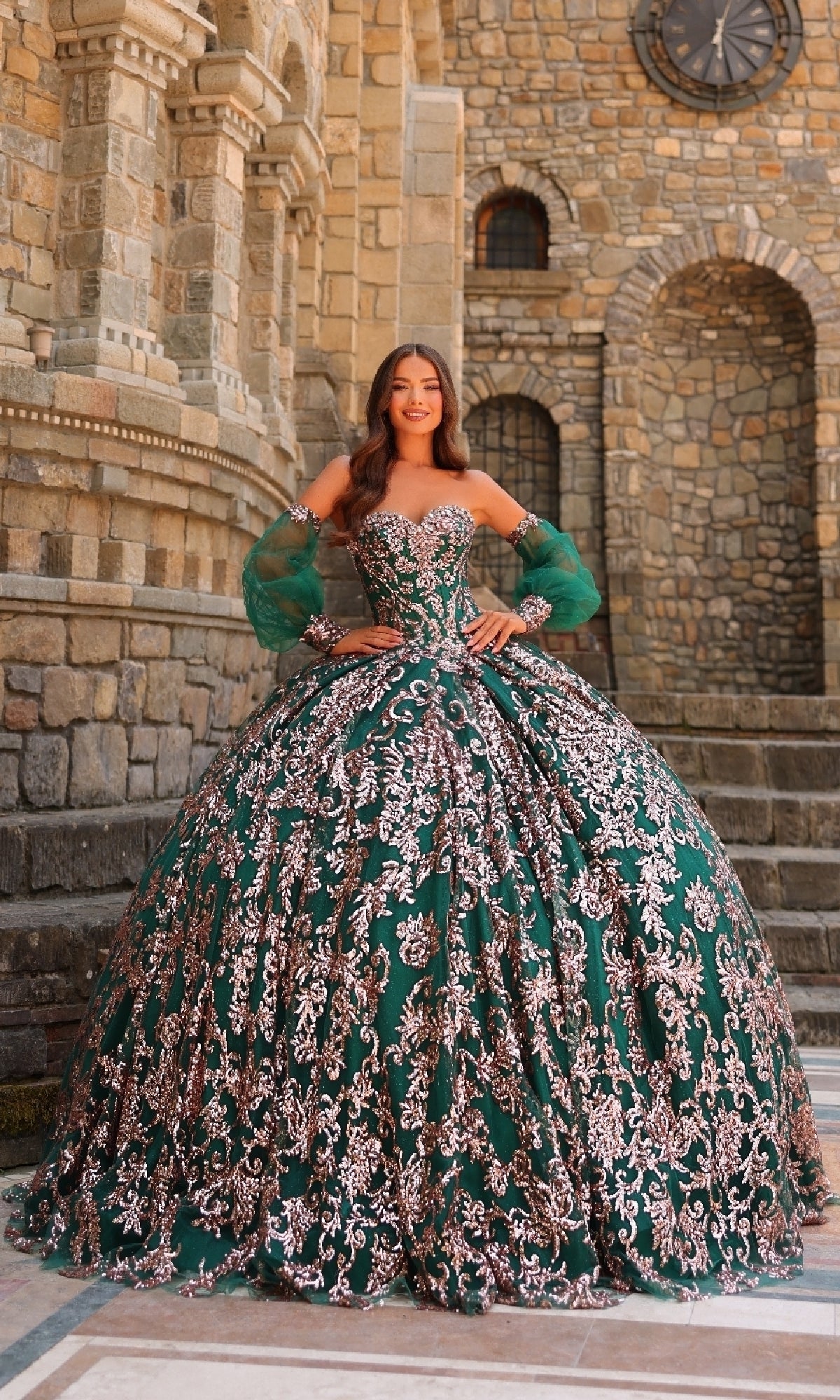 A woman stands outside a stone building wearing the Sequin Quinceanera Dress Amarra 54238, featuring a green ball gown style with intricate silver embroidery, a corset bodice, and sheer sleeves.